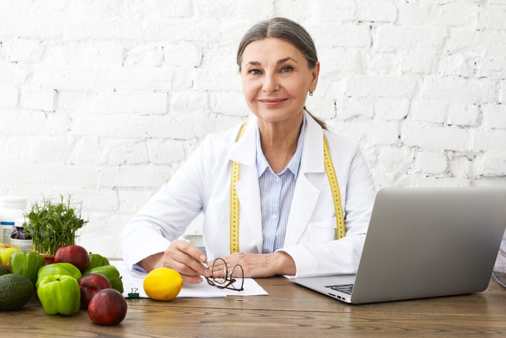 Indoor shot of positive smiling Caucasian female nutritionist in her sixties working online, sitting in front of open laptop pc, consulting clients on nutrition via social networks, making notes