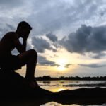 Un hombre estaba sentado con las rodillas dobladas y las manos sobre la cara en la base de un árbol y había agua alrededor.
